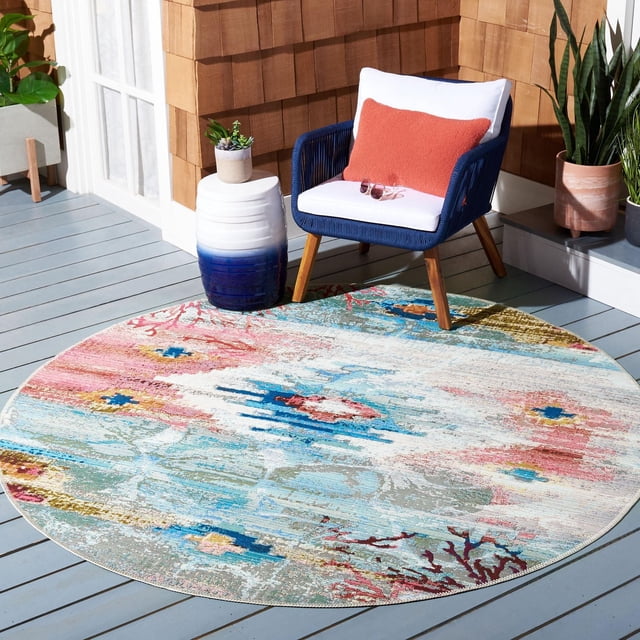 SAFAVIEH Barbados Aztec Corals Outdoor Area Rug, round with a multicolored abstract coral and ocean pattern in blue, pink, coral, and beige. Placed on a light gray wooden deck with a navy blue woven armchair featuring white cushions and a coral pillow, beside a white and blue ceramic side table with a small plant. Made from durable synthetic yarns, ideal for outdoor patios and coastal decor, also suitable for enhancing tropical outdoor living spaces.