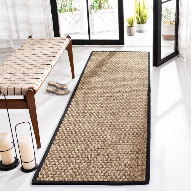 Safavieh Natural Fiber Arbor Border Area Rug runner with woven sisal texture and black border on white floor, beside a wooden bench with woven leather straps and glass doors with potted plants, suitable for coastal or casual hallway decor.