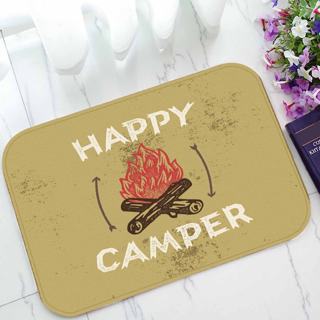 YUSDECOR Happy Camper Campfire Home Decor Floor Mat Area Rug Doormat, size 15.7x23.6 inches, featuring a beige background with a campfire graphic and the text 'Happy Camper' in bold white letters. Made with a neoprene rubber backing for durability and stain resistance, suitable for indoor use in bathrooms, bedrooms, or offices. Shown placed on a white tiled floor with flowers and a book nearby.