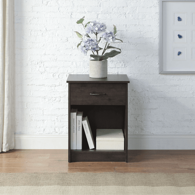 Mainstays table de Chevet à un Tiroir, Fini Chêne Foncé, avec un Tiroir et une Tablette Ouverte pour Tenir des Livres et une Boîte de Rangement, Surmontée d'Un vase de Fleurs Violet Pâle, Posée sur un Plancher de Bois contre un Mur de Briques Blanches, Convient comme table de Chevet Ou d'Appoint dans les Chambres d'Enfants.