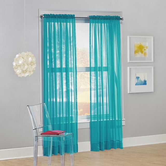 SUN TEXTILE VOILE ROD POCKET CURTAIN in teal sheer voile fabric with built-in rod pockets, hanging on a silver rod over a window in a modern room with light wood flooring, a clear acrylic chair holding books, and two framed abstract artworks on a gray wall, softly filtering light while enhancing privacy and suitable for layering.