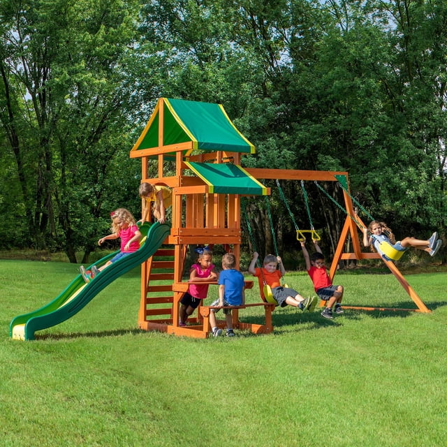 Backyard Discovery Tucson Swing Set with wooden frame and green fabric-covered upper deck and snack stand, featuring an 8-foot slide, rock wall ladder, two belt swings, trapeze bar, and picnic table below on a grassy lawn with children playing.