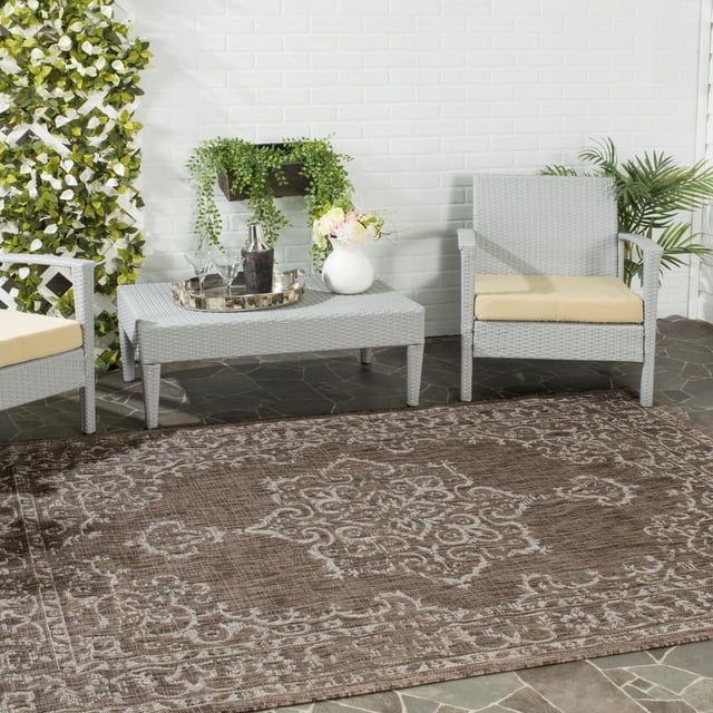 SAFAVIEH Courtyard Clodagh Medallion Indoor/Outdoor Area Rug in brown with detailed medallion pattern, placed on stone patio floor between two light gray wicker chairs with beige cushions and a matching wicker coffee table holding a silver tray and white vase with flowers, surrounded by green plants and white brick wall, ideal for durable indoor and outdoor use.