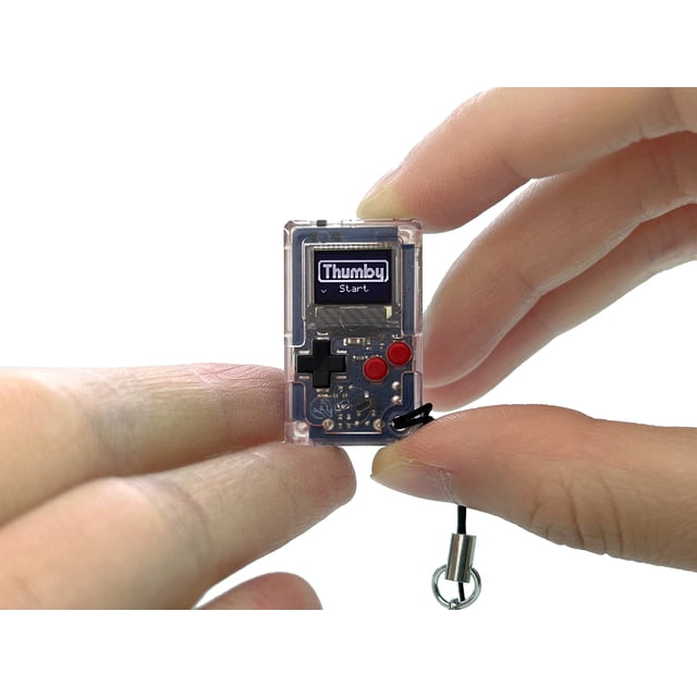 Tinycircuits Thumby (clear) tiny game console, a miniature playable and programmable keychain, shown held between fingers. Features a clear casing, six functional buttons, and a small screen displaying 'Thumby Start.' Preloaded with retro games, it includes a keychain attachment for portability and STEM learning applications.