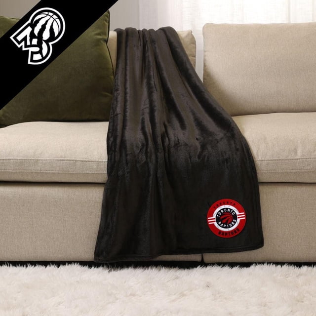 NHL, NBA, MLB Black Sports Throw Blanket 50" x 60" draped over a beige sofa with a green cushion, made of plush 100% polyester fabric, featuring a red and black Toronto Raptors logo near the bottom edge, suitable for indoor or outdoor use.