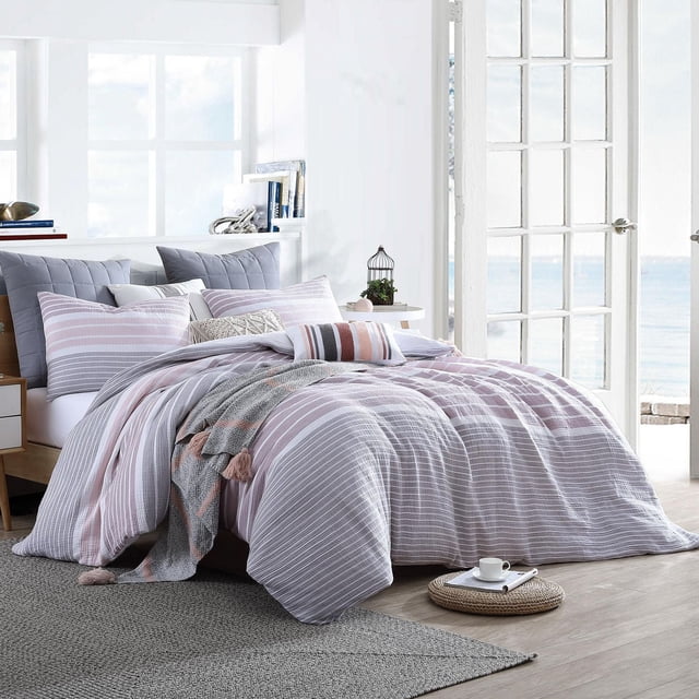 Ensemble de Housse de Couette en Coton – Collection Cordelia avec des Rayures Roses, Blanches et Grises Douces, des Taies d'Oreiller Assorties et une Couverture Jetée à Pompons sur un Lit dans une Pièce Lumineuse avec de Grandes Fenêtres et un Plancher de Bois Clair. l'Ensemble Est Lavable à la machine avec Fermeture à Boutons, Convient pour Ajouter du style et du Confort à Toute Chambre.