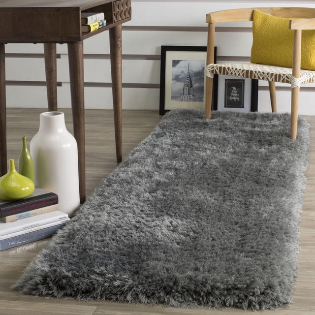 Safavieh Luxe Paxton Solid Plush Shag Area Rug, Grey, 2'3" x 8' runner featuring thick plush synthetic yarns for softness, placed on a light wood floor beside a wooden chair with a yellow cushion and a dark wood table with decorative vases and books, suitable for enhancing warmth and texture in contemporary home decor.