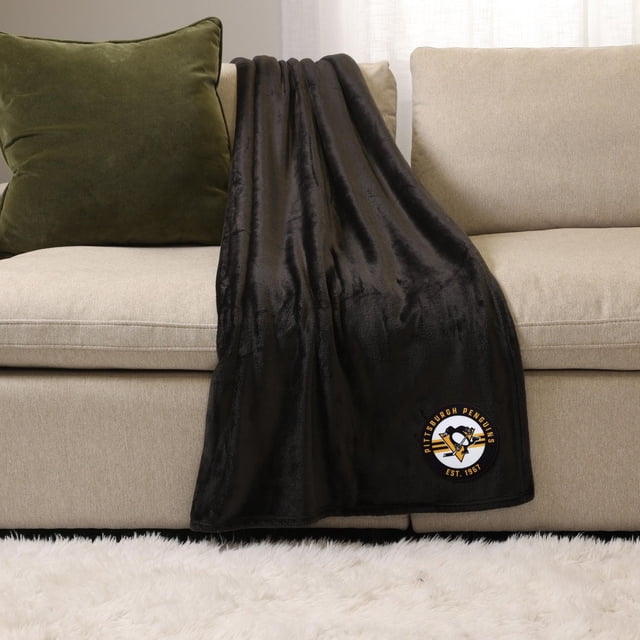 NHL, NBA, MLB Black Sports Throw Blanket, 50" x 60", draped over a beige sofa with a green cushion, made of soft plush 100% polyester with a Pittsburgh Penguins embroidered logo near the bottom edge, ideal for cozy indoor use and warmth.