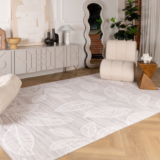 Rug Branch Modern Floral Indoor Area Rug in cream and light grey with a large leaf pattern, made of 100% polyester for easy cleaning, placed on a light wood herringbone floor in a modern living room with a beige boucle lounge chair, wooden side table, wavy full-length mirror, and grey sideboard with abstract decor, ideal for enhancing home interiors.