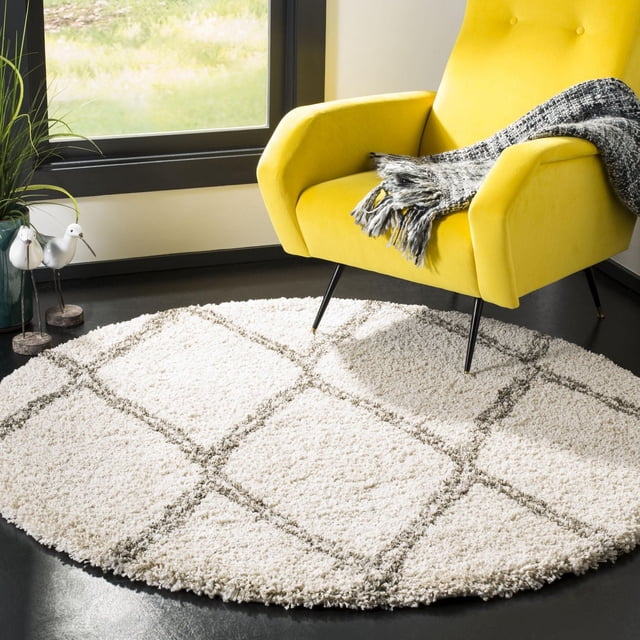 SAFAVIEH Hudson Adella Geometric Diamonds Shag Area Rug, round with a thick ivory shag pile and gray geometric diamond tile pattern, placed on a dark floor beside a yellow modern armchair with a gray throw, suitable for adding texture to contemporary living areas.