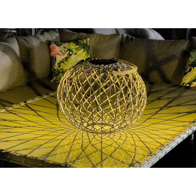 Dura Solar Rattan Effect Lantern with a spherical woven rattan frame on a yellow outdoor table casting intricate lattice shadows, ideal for patios and balconies, shown with cushioned seating and tropical leaf-patterned pillows in the background.