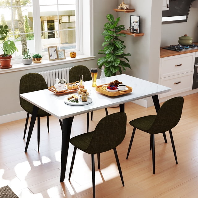 Homy Casa 63 inch modern dining set with a rectangular white faux marble tabletop and black steel legs, paired with four beige fabric upholstered chairs with cushioned seats and supportive backs, set in a bright kitchen with light wood flooring, breakfast trays with food and drinks on the table, and a green potted plant in the corner.
