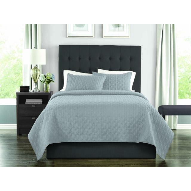 Springmaid 3 Piece Quilt Set, Solid Colours Easy Care in grey microfiber fabric on a bed with a black tufted headboard, featuring a textured quilt and matching pillow shams in a bedroom with dark hardwood floors, a nightstand with a lamp and flowers, and light-coloured curtains. The set is suitable for adding a calm, durable bedding layer.