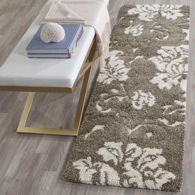 Safavieh Florida Merrick Floral Shag Area Rug displayed on a light wood floor next to a white bench with gold metal legs. The rug features a sculpted high-low pile with a crème tone-on-tone damask vine floral pattern, crafted from durable power-loomed synthetic yarns, suitable for enhancing home accent areas.