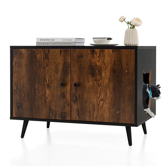 Gymax Industrial Cat Litter Box Enclosure Hidden Washroom Cat House End Table Cabinet with a black and natural wood finish, tapered metal legs, and a cat-shaped side entry for ventilation and easy access. The double-door cabinet hides the litter box and provides storage space for cat supplies. The tabletop is styled with books, a bowl, and a vase with flowers, making it suitable as a side table. A cat is seen exiting through the side entry.