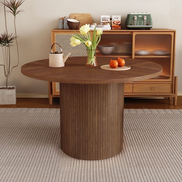 Wirrytor Dining Table, a modern farmhouse round kitchen table with a 47-inch walnut wood tabletop and a unique wood strip base. The table is styled in a cozy dining room setting with a light-coloured rug and a wooden sideboard in the background. Suitable for seating up to 4 people, this versatile table is ideal for kitchens, dining rooms, or living rooms.