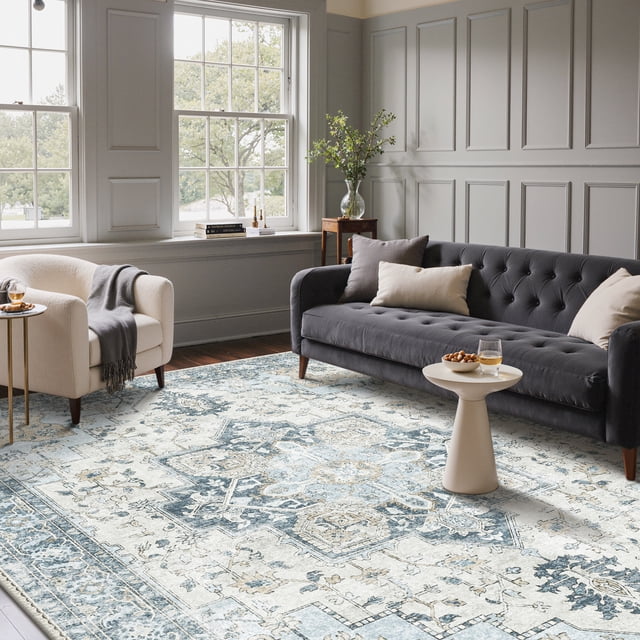 KUETH 9'x12' area rug displayed in a living room with a vintage-inspired blue and white design. The ultra-thin, machine washable rug lies flat under furniture, including a grey tufted sofa and beige armchair, complementing the modern decor. Designed for high-traffic areas, it features a durable woven polyester surface and non-slip rubber backing, ideal for living rooms, bedrooms, and dining rooms.