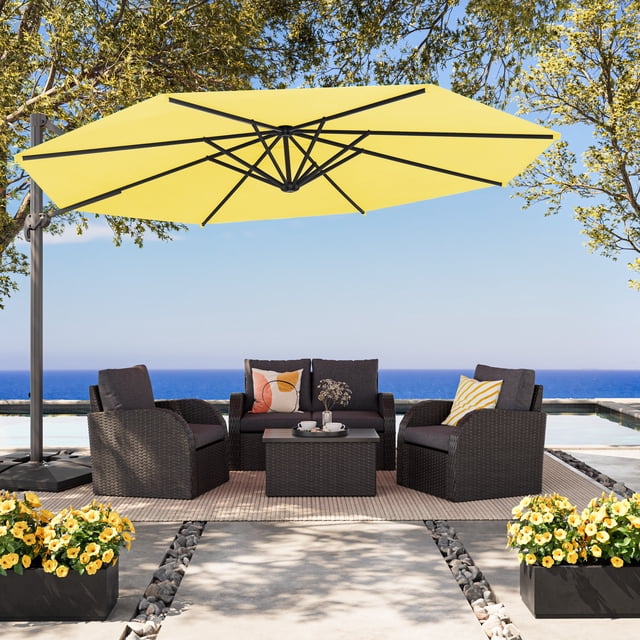 CorLiving 11.5ft Deluxe Tilting and Rotating Offset Patio Umbrella with Aluminum Frame and Durable Yellow Canopy, shading a dark grey woven outdoor seating set with cushions and decorative pillows on a beige rug beside a pool with ocean view, framed by trees and yellow flowering plants in black planters.