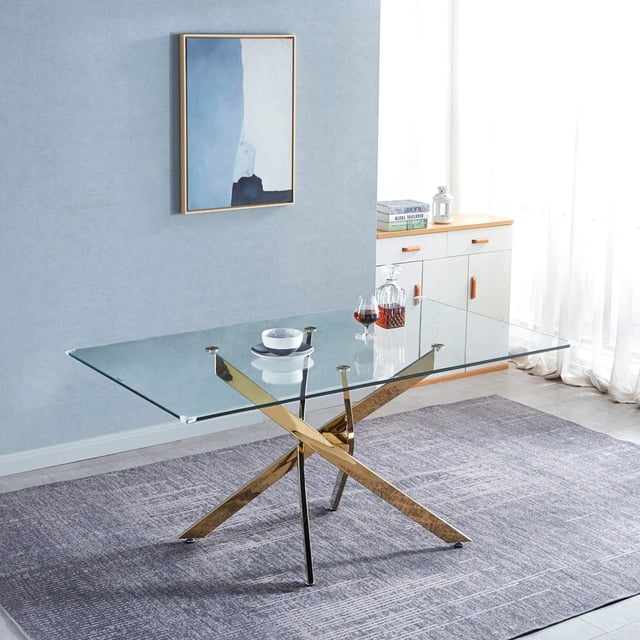 Heavenly Collection Gold Rectangular Tempered Glass Dining Table with Cross Metal Base featuring a clear tempered glass top supported by a gold metal X-frame base with felt foot pads to protect floors, set on a gray textured rug in a modern dining room with light blue walls, abstract wall art, and a white sideboard with wooden handles. This dining table is also suitable for use as a stylish workspace or craft table.