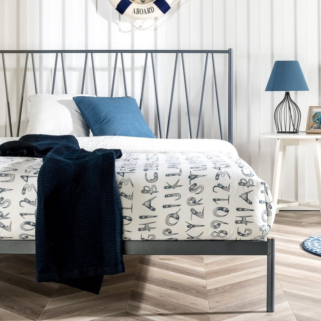 Zinus Olga Metal Platform Bed with Headboard in blue, twin size, featuring a geometric slatted headboard and navy blue steel framework with brass accents. The bed is shown in a modern bedroom setting with a white and navy alphabet-patterned bedding, a navy throw blanket, and a matching blue pillow. The platform bed eliminates the need for a box spring with its sturdy metal slats, suitable for various mattress types. A white side table with a blue lamp and nautical decor completes the scene.