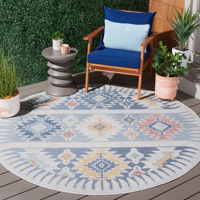SAFAVIEH Sarasota Giovanni Tapis d'Aire Intérieur/extérieur Aztèque, Rond avec un Motif Aztèque Multicolore dans des Tons Bleu, Jaune et Rouge, Placé sur un Plancher de Terrasse Extérieur Gris. Il Est Styleé avec un Fauteuil en Bois avec des Coussins Bleus, une table d'Appoint Sculpturale Grise Tenant une Tasse Rose et une Petite Plante, et Entouré de Plantes en Pot Près d'Un Mur de Haie Verte, Convient à une Utilisation Intérieure et Extérieure.