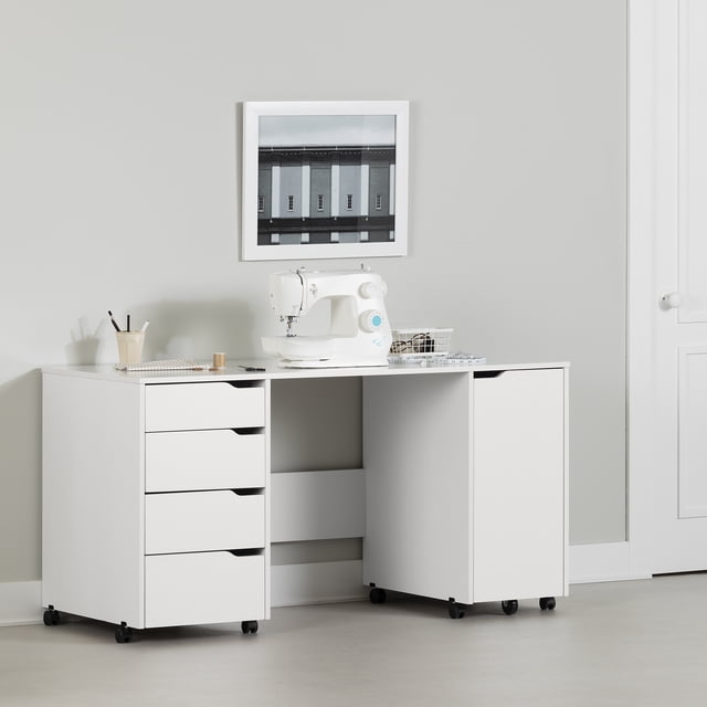 South Shore, Crea Sewing Craft Table on Wheels in pure white with a scratch-resistant top surface holding a white sewing machine, four drawers on the left, and a sliding storage module on the right with casters and cut-out handles, ideal for sewing and crafts, shown in a neutral room with a framed black and white photo above.
