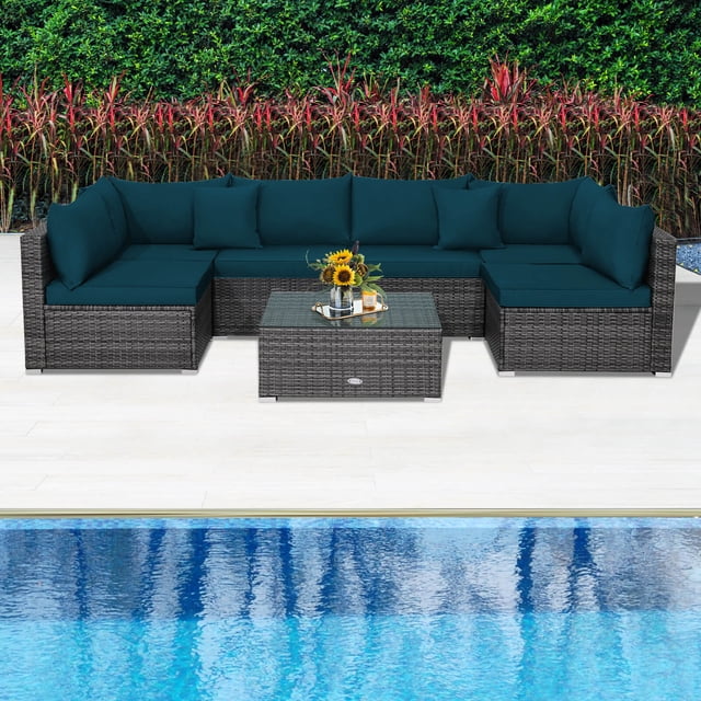 Costway 7PCS Patio Rattan Furniture Set Sectional Sofa Cushioned Garden in Peacock Blue. This outdoor sectional set includes a mix grey PE rattan frame with weather-resistant properties, featuring 2 corner sofas, 4 armless sofas, and a tempered glass coffee table. The cushions are thickly padded in peacock blue with removable covers for easy cleaning. Includes 2 throw pillows for added comfort. The modular design allows for flexible arrangements, suitable for patios, gardens, or poolside settings. The set is shown placed on a light tile patio near a pool, with a lush green hedge and red foliage in the background.