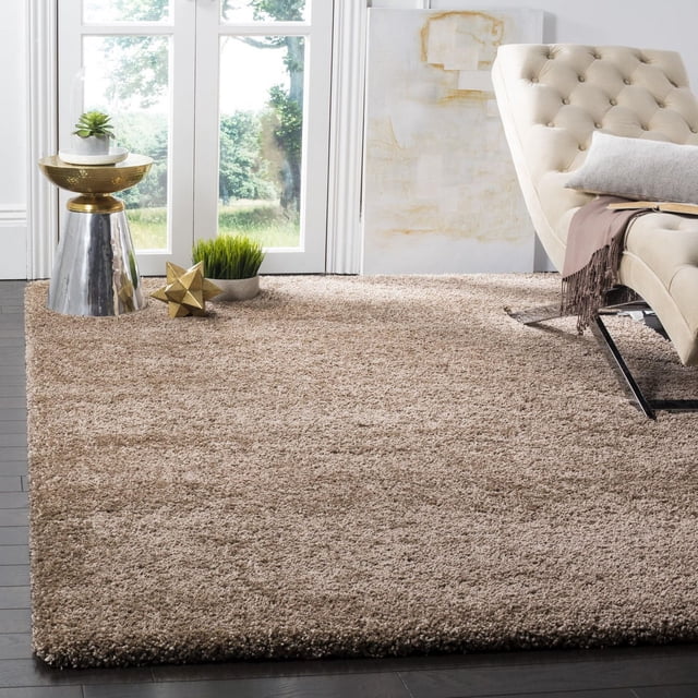 Safavieh Milan Harlow Tapis Shag Uni en beige, Montré dans un Salon Lumineux avec des Planchers de Bois Franc Sombres. le Tapis Shag Moelleux Couvre le Sol sous un sofa Tufté Crème avec des Pattes en Métal, Accentué d'Un Coussin Gris et d'Un Jeté Brun. À Proximité Se Tient une table d'Appoint Métallique avec des Finitions or et Argent Tenant une Petite Succulente en Pot, à Côté d'Une sculpture Géométrique Dorée et une Plante Verte. une Grande Peinture Abstraite Repose contre un Mur Blanc Près de Portes en Verre Montrant la Verdure Extérieure. Ce Tapis en Fil Synthétique durable Ajoute