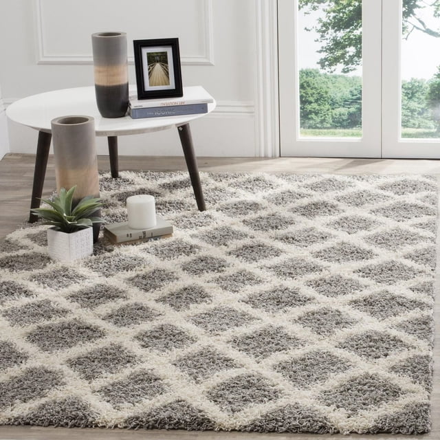Safavieh Layla Geometric Plush Shag Area Rug featuring a grey and cream diamond pattern with thick plush synthetic fibers, displayed in a sunlit room with hardwood floors and large glass doors, beneath a white round table with dark wooden legs holding decorative vases, books, a candle, and a small plant, ideal for high-traffic home areas.