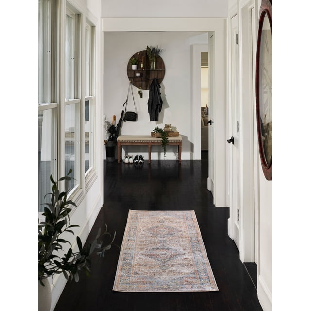 ECARPET Irina Traditional Runner Rug with muted Turkish-inspired patterns in soft blues, greys, and rust tones on dark hardwood floor in a white hallway featuring a beige cushioned bench with shoes underneath, a round wooden wall shelf with plants, and natural light from large windows. Suitable for high-traffic areas like entrances or hallways.