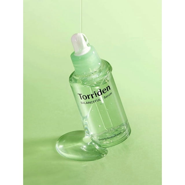 Torriden Balanceful Cica Serum 50ml clear glass bottle tilted with watery serum dripping from the dropper onto a pale green surface, highlighting its lightweight, soothing formula with Centella Asiatica extract to calm sensitive and acne-prone skin and reduce redness.