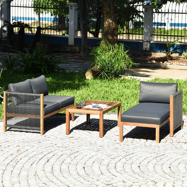 Gymax 3PCS Patio Acacia Wood Sofa Furniture Set with loveseat, lounge chair, and coffee table. Made of sturdy acacia wood, it features nylon rope armrests and thick, dark cushions for comfort. Ideal as a patio conversation set, it is displayed on a stone patio surrounded by lush greenery.