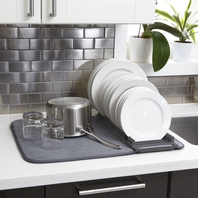 Umbra UDry Dish Drying Mat in charcoal with a removable dish rack holding white plates on a white kitchen countertop with stainless steel tile backsplash. The microfiber mat absorbs water and protects surfaces, also holding upside-down clear glasses and a stainless steel saucepan. The mat folds compactly for storage and is machine washable, suitable for organized dish drying and countertop protection.