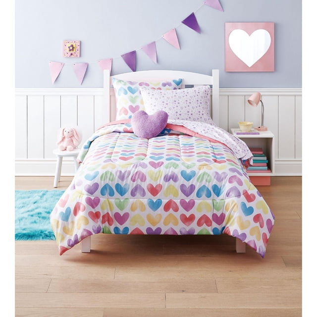 Mainstays Kids Hearts Bed in a Bag twin comforter set with pastel multicolored heart patterns on soft microfiber fabric. The reversible comforter shows a white side with small purple hearts. Includes matching pillowcases, fitted and flat sheets, and a purple heart-shaped decorative pillow. Styled on a white wooden bed frame in a child's room with light wood flooring, a pink and purple pennant banner, a white nightstand with a pink lamp, and a plush pink bunny on a stool. Machine washable and suitable for all seasons.