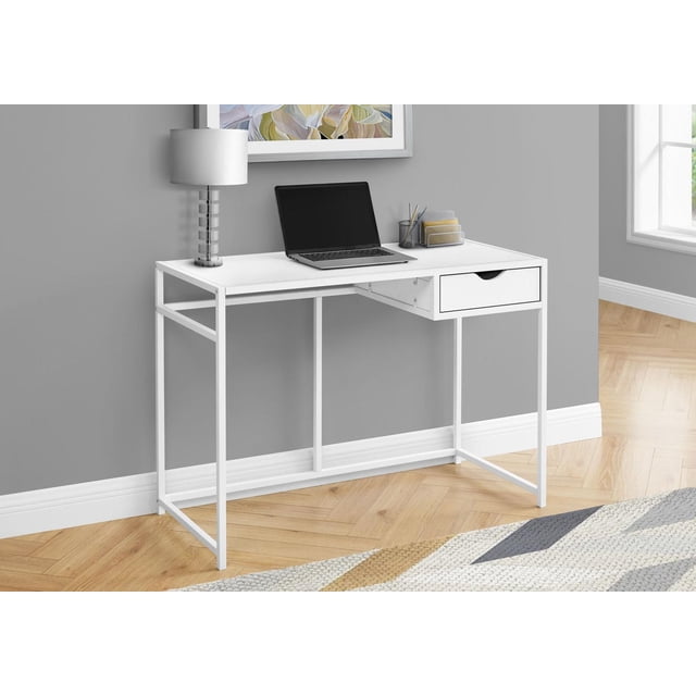 Monarch Specialties computer desk with white laminate top and slender white metal frame, featuring one storage drawer with cutout handle and metal slides, ideal for compact home office spaces, shown with a laptop, lamp, and office supplies on a light wood herringbone floor and gray wall background.