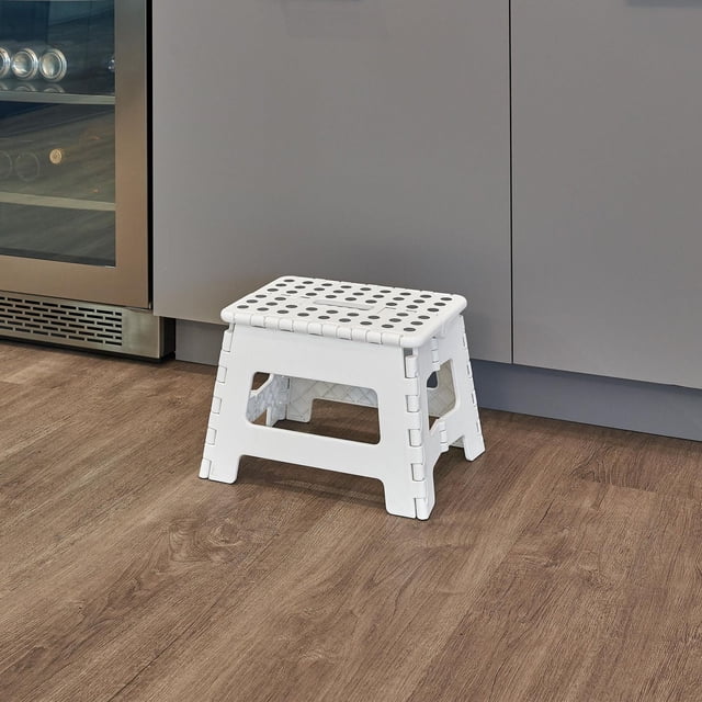 MAINSTAYS Foldable Step Stool in white plastic with perforated top, unfolded on wood floor by grey kitchen cabinets and stainless steel wine cooler, sturdy and space-saving for laundry rooms, bathrooms, closets, or garages, also suitable as a compact seat or footrest.
