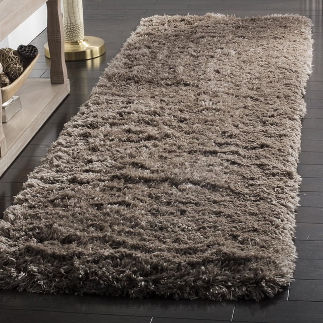 SAFAVIEH Polar Abbot Solid Extreme Plush Shag Area Rug in taupe with a 3-inch thick plush pile, displayed on dark hardwood flooring beside a light wood console table and brass lamp base, suitable for adding warmth and soft texture to runner rug areas.