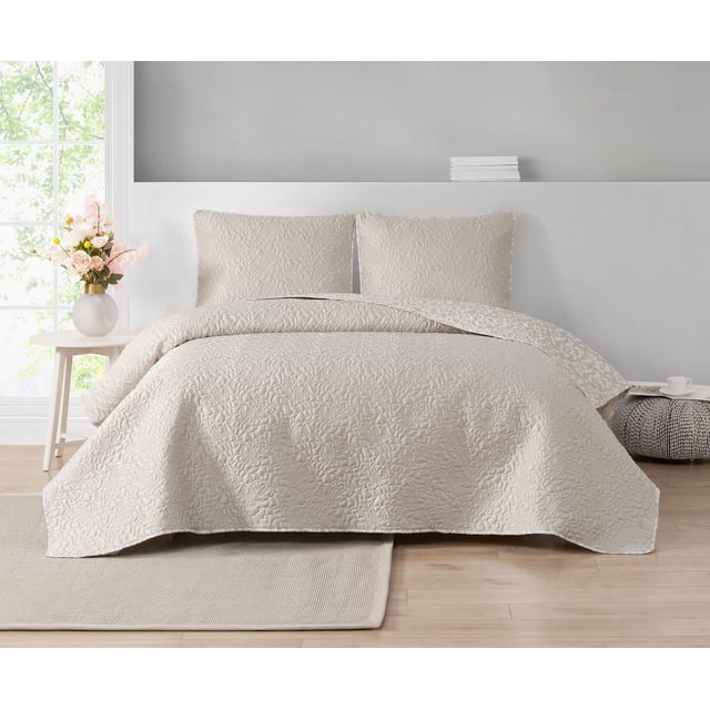 Valerie 3 Piece Reversible Quilt Set on a bed with two matching shams, featuring stitched large flower outlines on a soft matte satin solid beige surface. The quilt is flipped to show a white floral botanical pattern on the soft microfiber reverse. The room includes light wood flooring, a beige rug, a side table with a flower vase, and a knitted pouf with a cup and saucer.