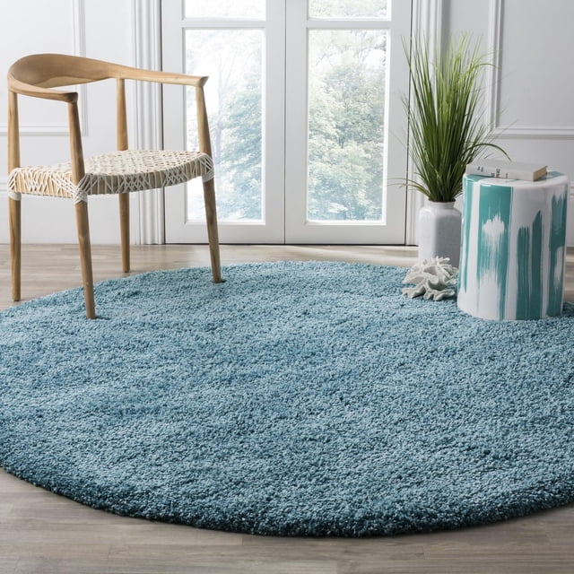 Safavieh California Solid Plush Shag Area Rug in round shape with a dense 4.5kg/m² polypropylene pile in blue, placed on light wood flooring in a bright room with a wooden chair with woven seat, a white and teal ceramic side table, and a tall green plant by glass doors, ideal for adding softness and warmth to living spaces.