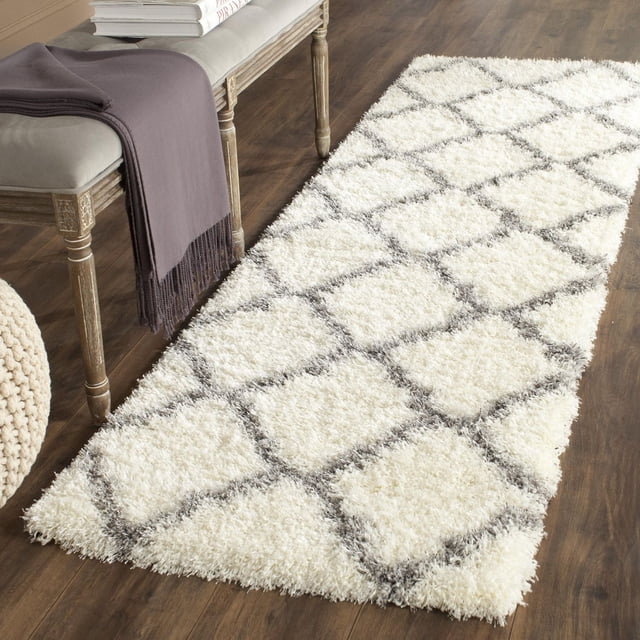 Safavieh Montreal Cebrail Geometric Shag Area Rug runner with a plush white shag pile and gray diamond geometric pattern, placed on dark hardwood flooring beside a wooden bench with a gray cushion and folded purple throw, suitable for contemporary or transitional interiors and often used to add warmth and texture to hallways or entryways.