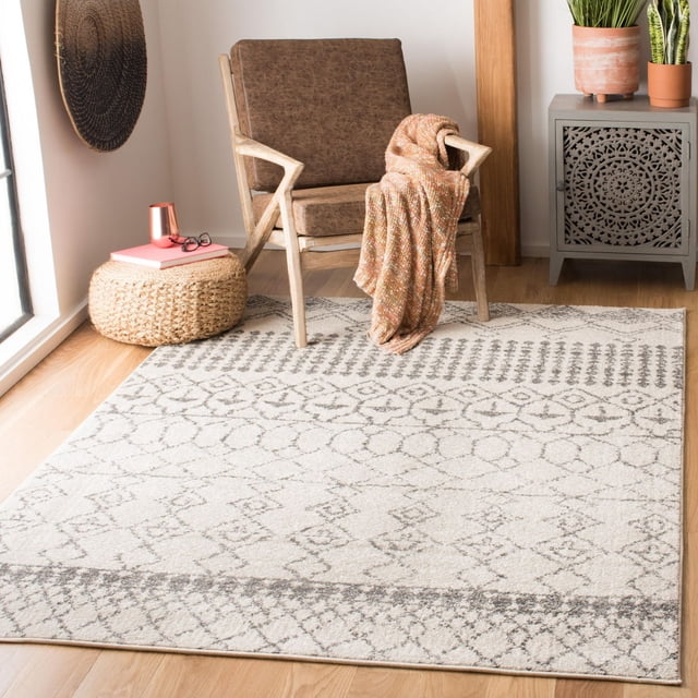 Safavieh Tulum Coriander Moroccan Geometric Area Rug in light neutral shades with gray geometric patterns, placed on a light hardwood floor under a brown cushioned wooden armchair draped with an orange throw, beside a woven ottoman with books and glasses, and a gray carved cabinet with potted plants, providing a plush texture and warm accent to contemporary décor.
