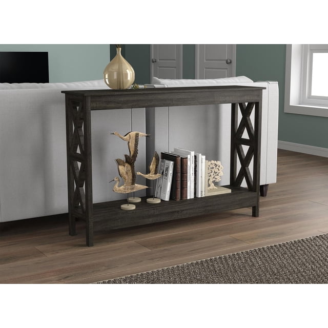 Safdie & Co. Console Table 47.25L Dark Grey 1 Shelf with open X-shaped side panels and dark grey wood finish, positioned behind a light grey sofa on wood flooring. The lower shelf holds wooden bird sculptures, books, and a coral ornament. This contemporary accent table provides extra storage and complements living room furniture.