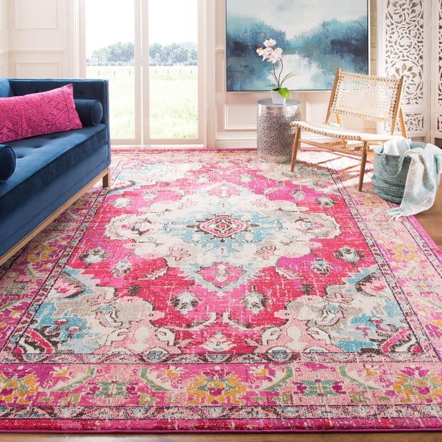 Safavieh Monaco Toria Traditional Area Rug with vibrant pink, blue, and cream Persian-inspired patterns, power-loomed polypropylene with mixed loop pile, displayed in a bright living room with a blue velvet sofa, woven chair, silver side table with orchid, and decorative white screen, ideal for adding Bohemian-chic flair to home interiors.
