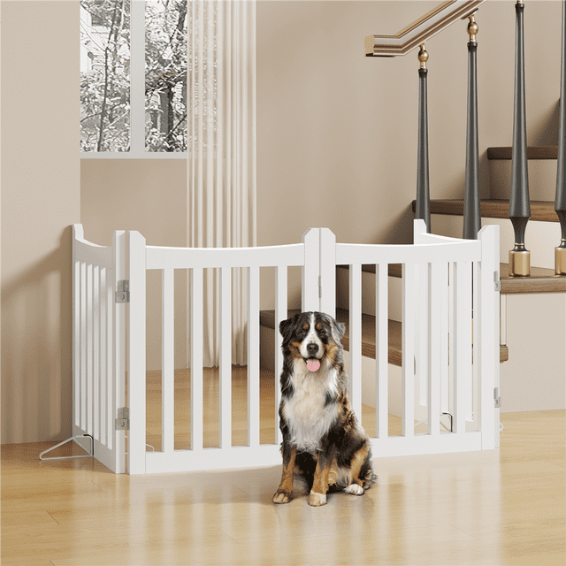 Yaheetech 4 Panel Freestanding Wooden Pet Puppy Fence with 2 Support Feet in white, displayed in a modern living room. The gate is made of durable MDF with five slats per panel, ensuring airflow and visibility. It is shown in a U-shaped configuration, emphasizing its foldable and adjustable design. A medium-sized dog is seated in front of the gate, illustrating its function as a pet barrier. The gate includes two metal support feet for stability and measures 79.5 inches long and 24 inches high when fully extended.