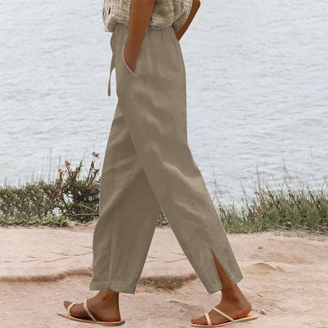 Women Casual Cotton Linen Drawstring Elastic Waist Long Wide Leg Pants in a solid beige color, featuring a middle-rise elastic waistband with a drawstring and wide-leg design. The pants are styled with a subtle side slit at the hem, suitable for casual outings or seaside holidays. Made from a blend of 60% cotton and 40% linen, offering comfort and breathability. The image shows a side view of the pants worn with sandals near a beach setting.