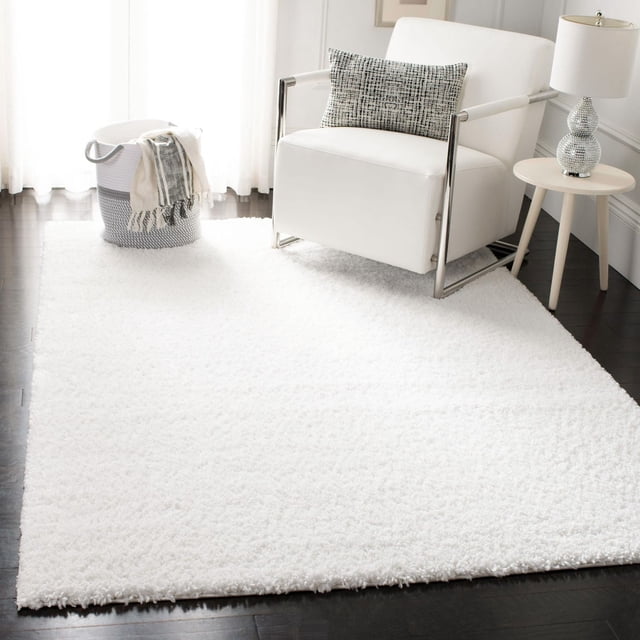 SAFAVIEH August Carlene Solid Plush Shag Area Rug in white, displayed on dark hardwood floors in a bright room, featuring a thick plush polypropylene texture under a modern white armchair with metal frame and patterned lumbar pillow, next to a round side table with a textured lamp and a woven basket with a throw blanket, ideal for adding softness and warmth to living areas.