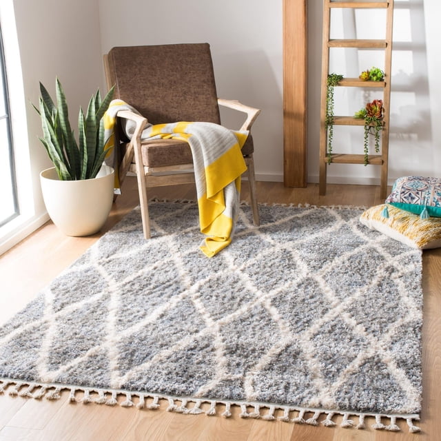 SAFAVIEH Berber Fringe Hattie Tapis Shag Géométrique avec Poil de 1,25 Po, Présentant un Motif Géométrique Losange Gris et Blanc et des Bords à Franges, Posé sur un Plancher de Bois Franc Clair sous un Fauteuil en Bois Brun Drapé d'Un Jeté Jaune et Gris, à Côté d'Une Plante Serpent en Pot et de Coussins Décoratifs, Convient pour Rehausser la Déco Maison boho-chic.