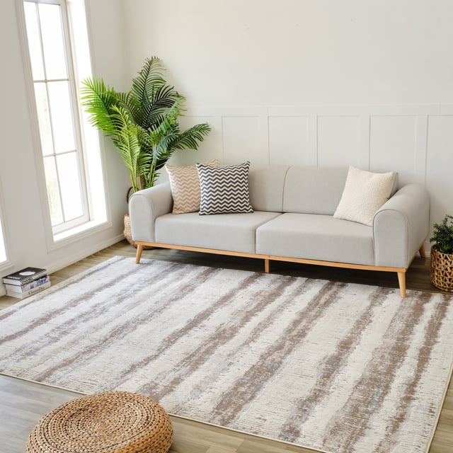 Rug Branch Nomad Abstract Indoor Area Rug in beige and brown, 7'x10' (6'7" x 9'6"), shown in a bright living room with a modern light grey sofa with wooden legs and three patterned cushions, a tall green plant near a large window, and a woven round floor cushion on light wood flooring, ideal for living room, bedroom, dining room, or kitchen.