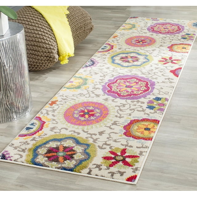 Safavieh Monaco Savannah Floral Area Rug 2'2"x8' runner with a cream background featuring vibrant circular floral patterns in pink, yellow, blue, and red, made of durable polypropylene power-loomed pile, placed on light wood flooring beside a knitted brown pouf with a yellow throw and a silver textured side table, often used as a bohemian-chic accent rug in home interiors.