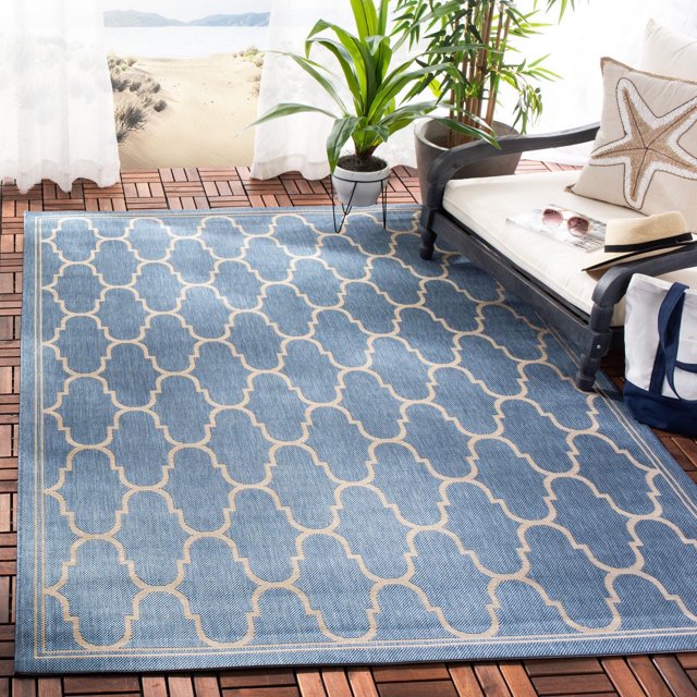 SAFAVIEH Courtyard Eustacia Tapis Géométrique Intérieur/extérieur en Bleu avec Motif Géométrique beige, Fabriqué en Polypropylène durable avec Tissage en sisal, Placé sur une Terrasse Extérieure en Bois. le Tapis Convient aux Espaces de Vie Occupés à l'Intérieur et à l'Extérieur. la Scène Comprend un Banc en Bois Foncé Rembourré avec un Coussin Étoile de Mer, une Plante en Pot et une Plage Sablonneuse visible à Travers des Rideaux Blancs Transparents.
